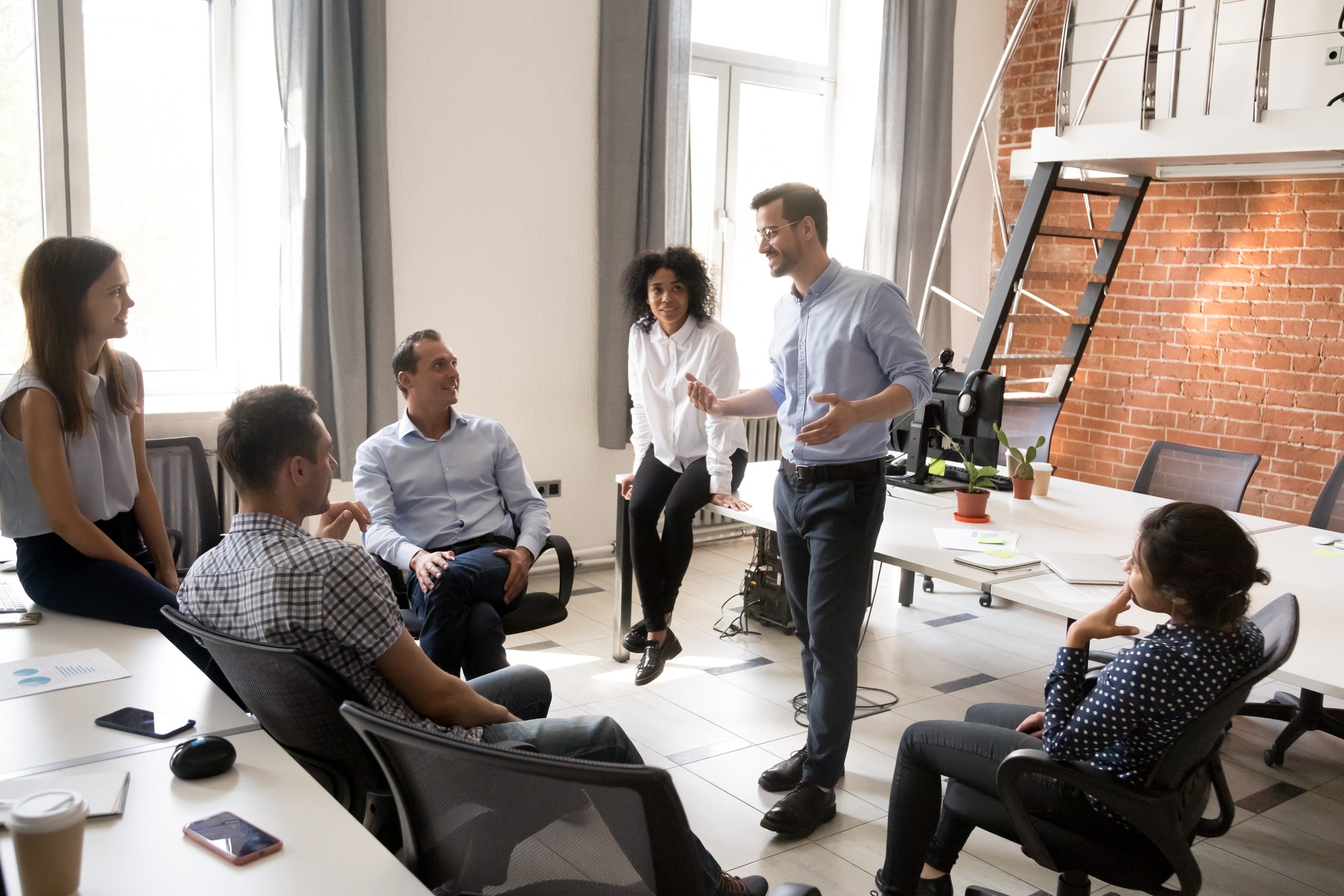 A diverse team engages in a productive discussion during a meeting in a modern office space with large windows and exposed brick walls.
