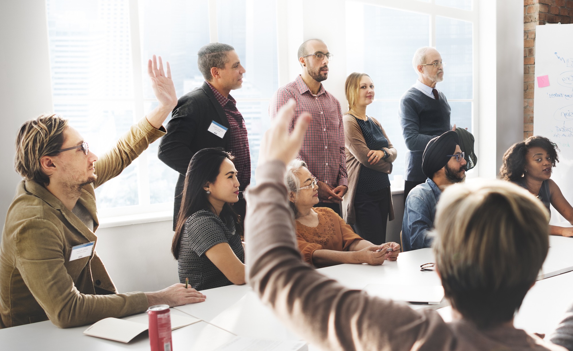 A diverse group of professionals engages in a meeting, with one person raising their hand to contribute. Natural light fills the room, fostering collaboration.