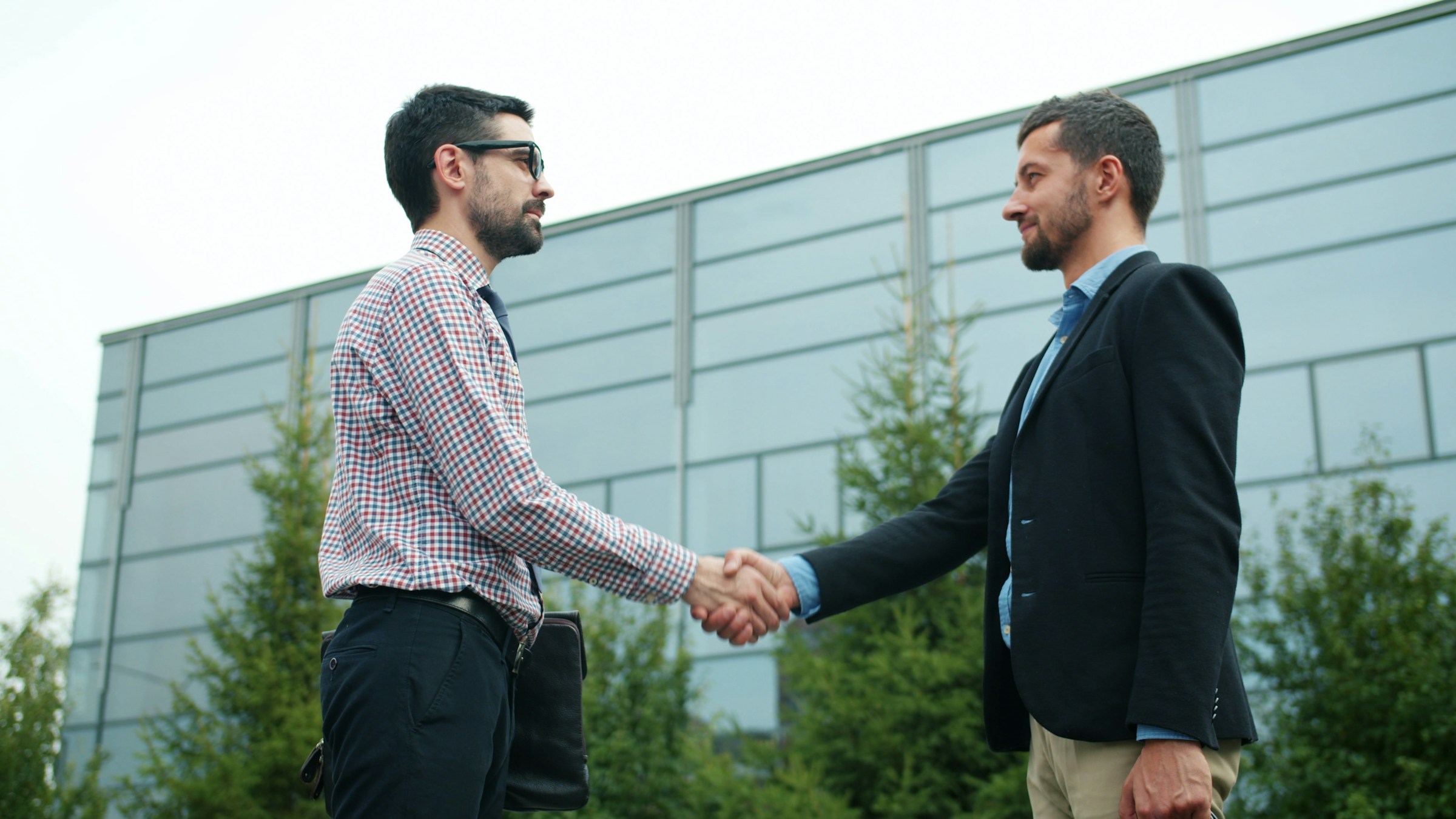 Two men shake hands outdoors, smiling, in front of a modern building. This image represents a professional greeting or business agreement.