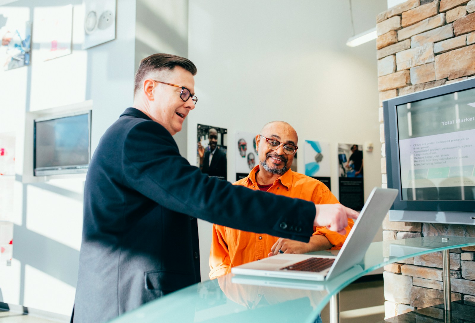 A professional man in a suit is engaged in a discussion with a smiling man in an orange shirt, pointing at a laptop together in a modern office setting.