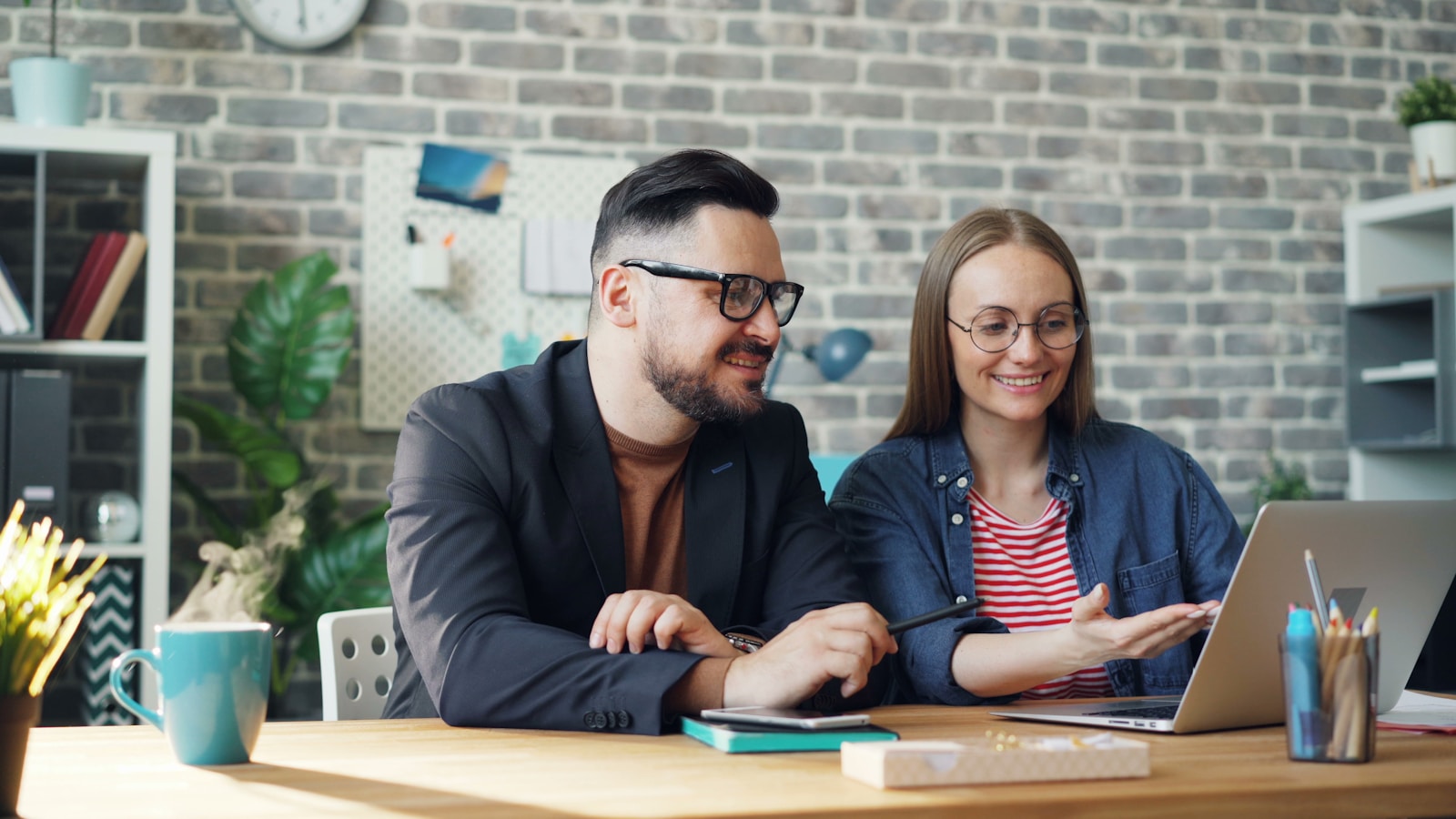 Two professionals collaborate at a desk, smiling and discussing ideas while using a laptop. The modern workspace features plants and creative materials.