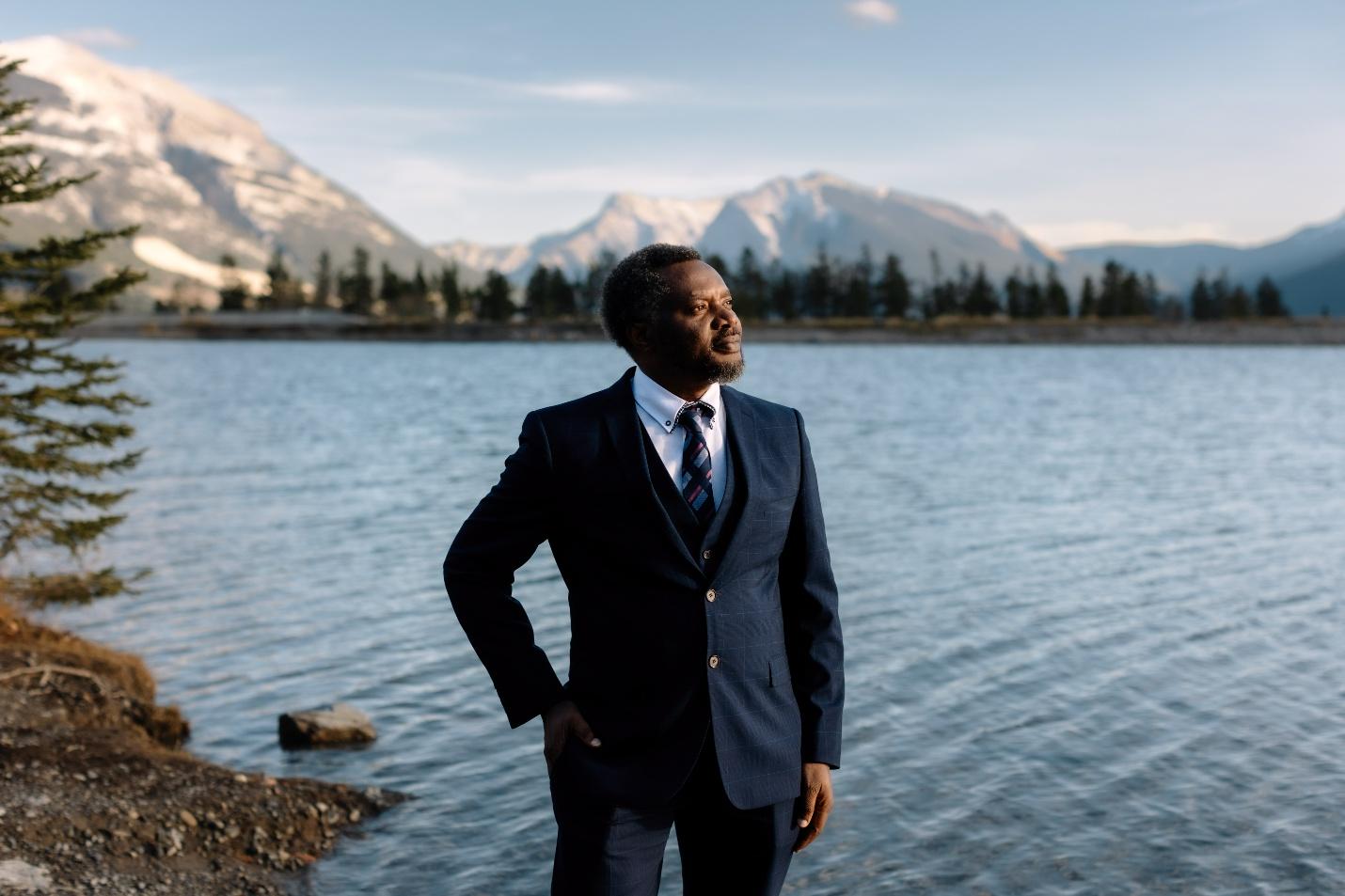 A man in a navy suit stands by a lake, gazing thoughtfully towards the mountains under a clear sky. The serene landscape underscores the moment's introspective nature.