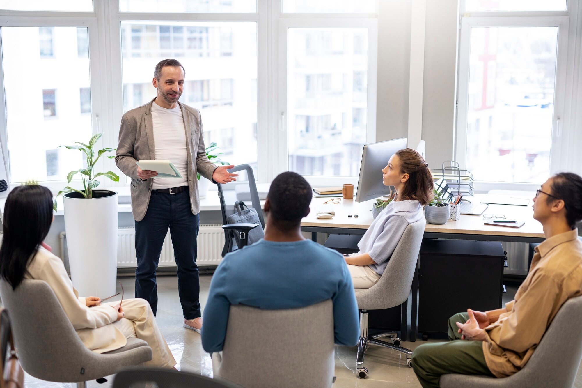 A speaker presents to a group in a bright office, fostering collaboration and discussion among the attendees. Plants and office supplies are visible.