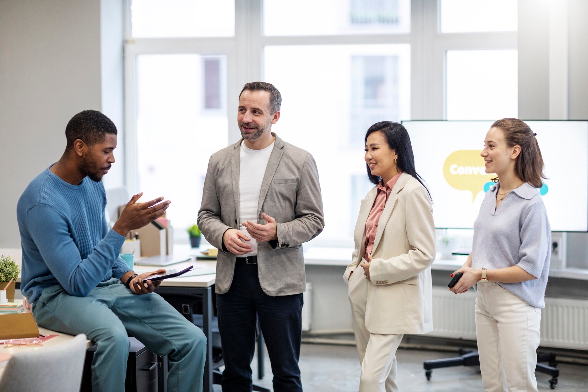 A diverse group of four people attentively listen to a speaker during a training session in a bright office, with a whiteboard displaying irregular verbs in the background.