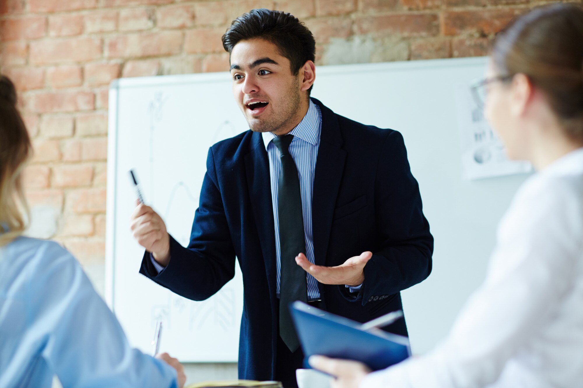 A young man in a suit passionately discusses a topic in a meeting, gesturing with a marker while engaging with colleagues.