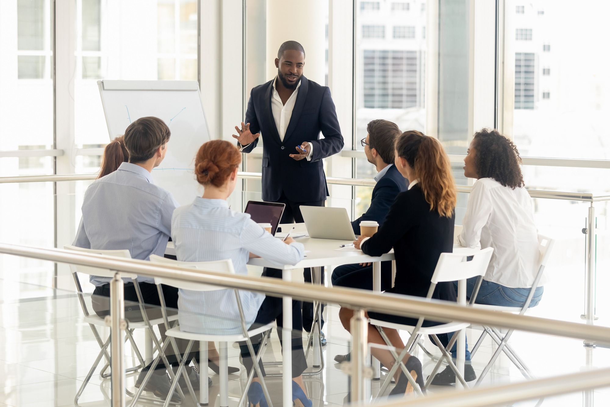 Smiling african American businessman stand make flip chart presentation for diverse work team in office, black male coach or trainer talk present strategy on whiteboard, training employees at meeting; Shutterstock ID 1389051551; Purchase Order: 9184412; Job: ; Client/Licensee: ; Other: